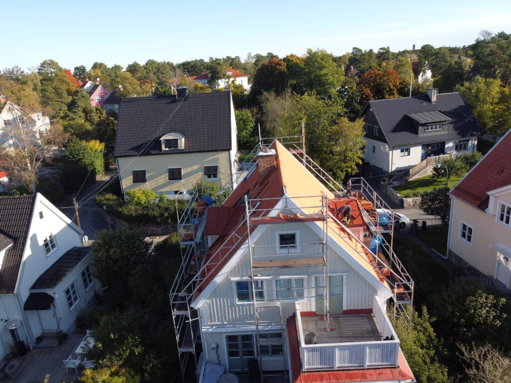 Fågelvy över hustak under takomläggning. Runt huset står flera byggställningar. I bakgrunden syns närliggande grannhus.