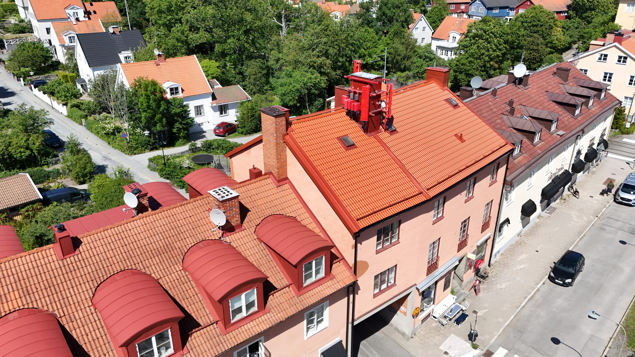 Fågelvy över en länga av lägenhetshus. Husens tak skiljer sig åt i färg och material. Bakom lägenhetshusen finns ett stort villaområde och stora träd.