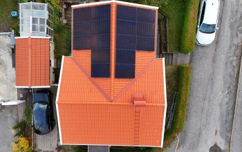 Fågelvy över hustak efter takomläggning. Hustaket är av tegelpannor, och har solceller, en takstege samt en skorsten i plåt. Runt huset syns delar av hustomten, samt två parkerade bilar.