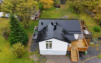 Fågelvy över ett vitt hus med svart, bandtäckt plåttak. I bakgrunden syns hustomten.