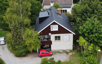 Fågelvy över ett stor hus i tegel och trä. Hustaket lutar och är gjort av svarta takpannor. Intill huset står en röd bil parkerad.