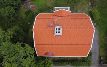 Fågelvy ovanifrån en stor villa med ett lutande tak och orangea tegelpannor. Taket har en skorsten, snörasskydd samt ett vindsfönster.