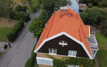 Ett stort villahus i trä och tegel, med sluttande tak och orangea takpannor. Taket har en skorsten, snörasskydd samt ett vindsfönster.